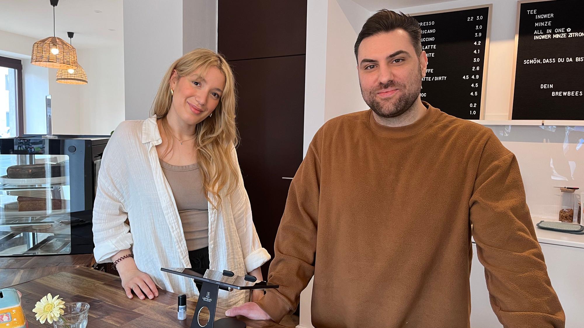 Eine Frau mit blondem Haar und ein Mann mit kurzen dunklen Haaren stehen hinter der Theke in einem Café.