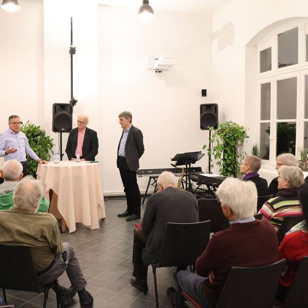 Drei Männer stehen bei einem Vortrag vor Publikum an einem Stehtisch.