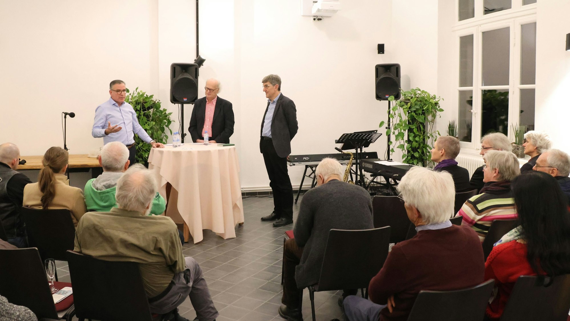 Drei Männer stehen bei einem Vortrag vor Publikum an einem Stehtisch.