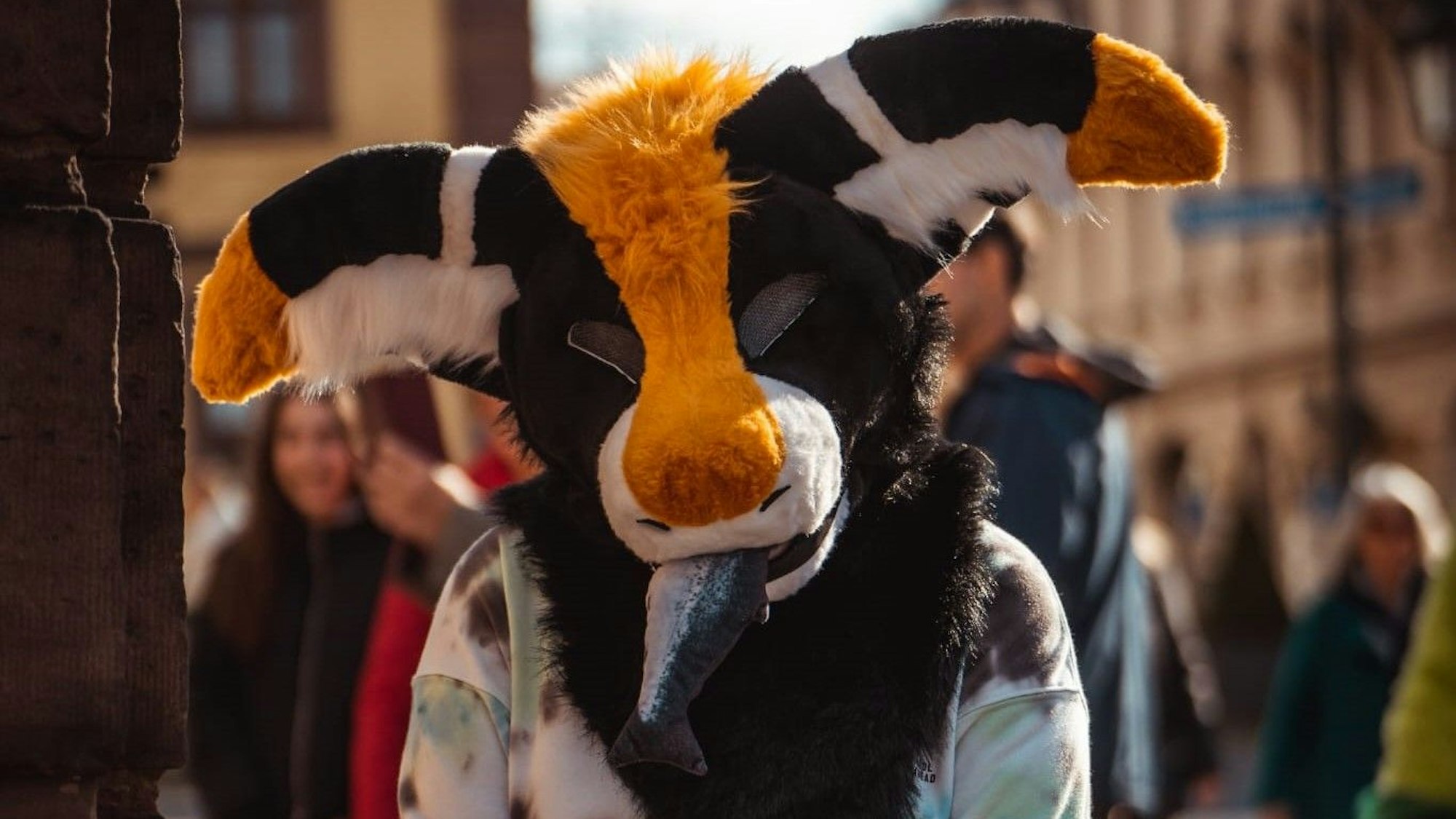 Eine 19-Jährige steht in einer Fußgängerzone und trägt eine plüschige Maske. Im Maul befindet sich ein Fisch aus Stoff.