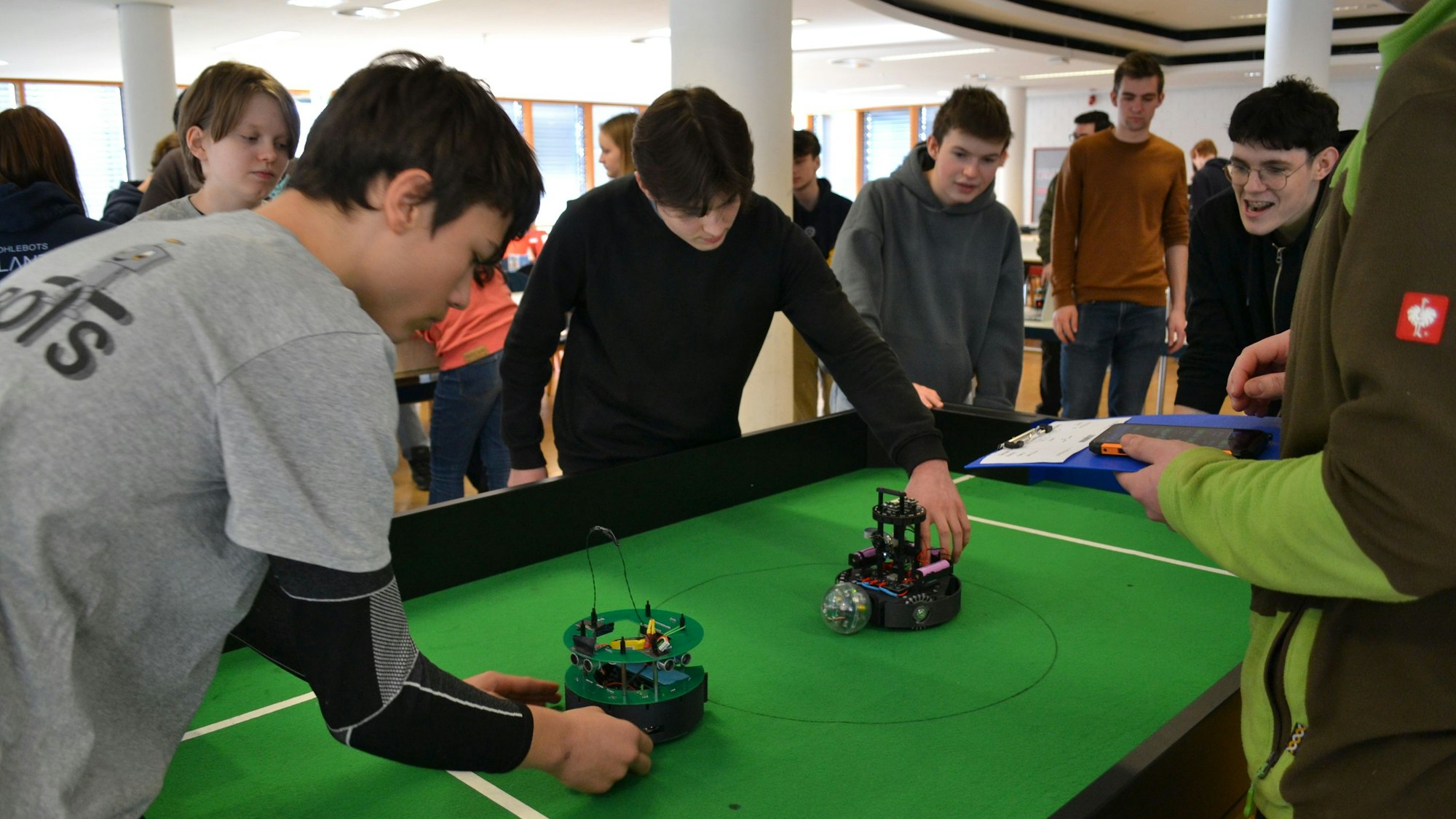 Jugendliche an einem Tisch, auf dem ein grünes Fußballfeld markiert ist. Sie starten kleine fahrbare Roboter.