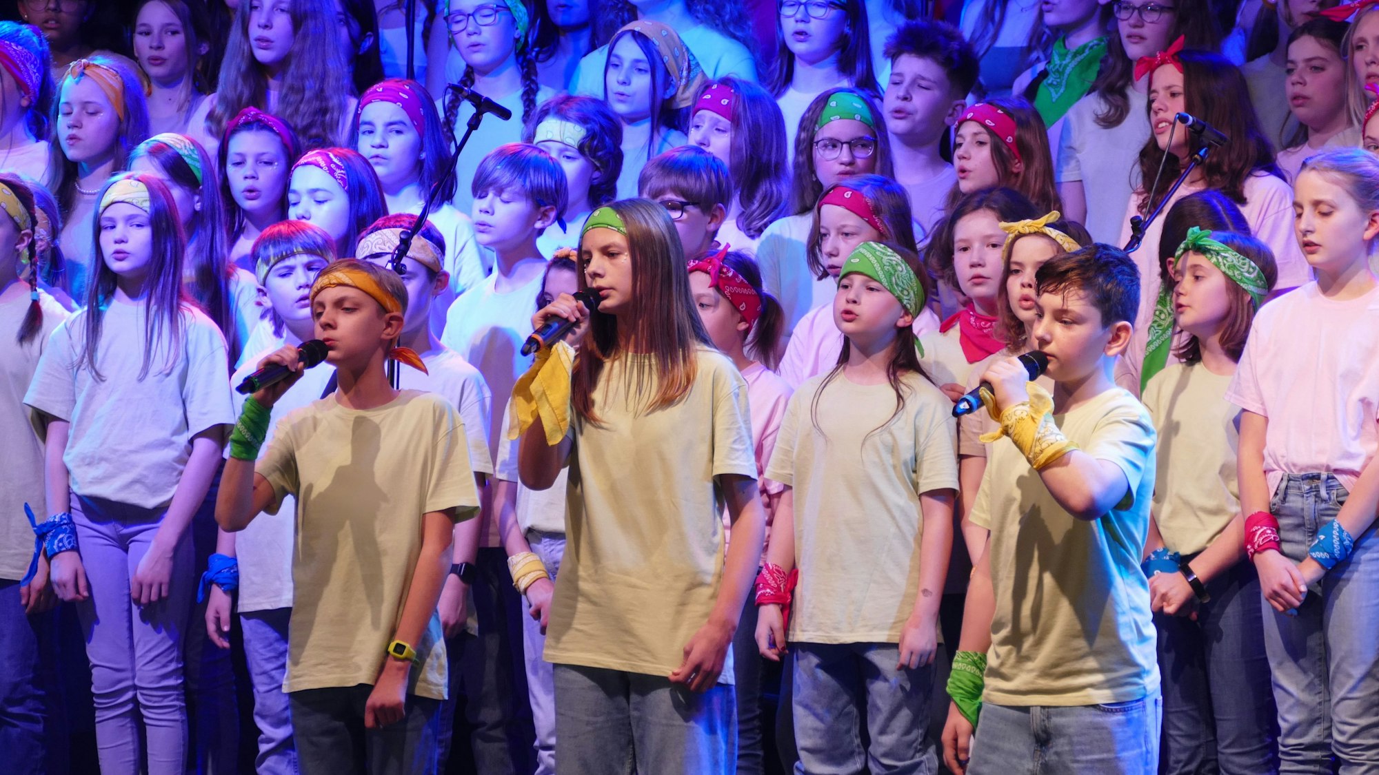 Ein Chor und vier Solisten singen auf einer Bühne. Sie tragen weiße T-Shirts, Jeans und bunte Haarbänder.