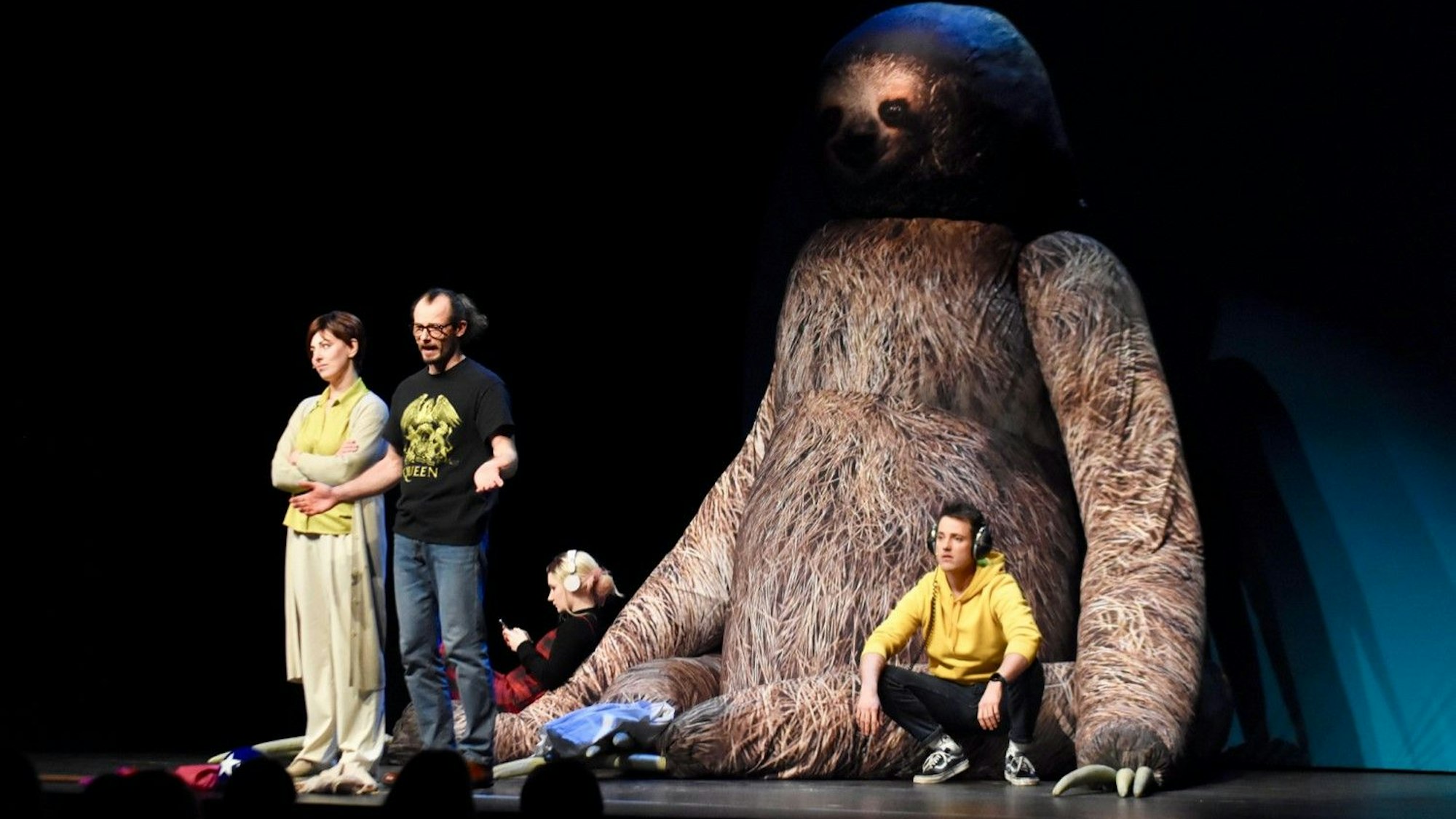 Szene aus dem Theaterstück Das „Das Pubertier“ in der Hennefer Halle Meiersheide: Eine Frau und ein Mann stehen auf einer Bühne, hinter ihnen sitzt ein riesengroßes Faultier, an das sich Schauspieler mit Kopfhörern und Handy anlehnen.