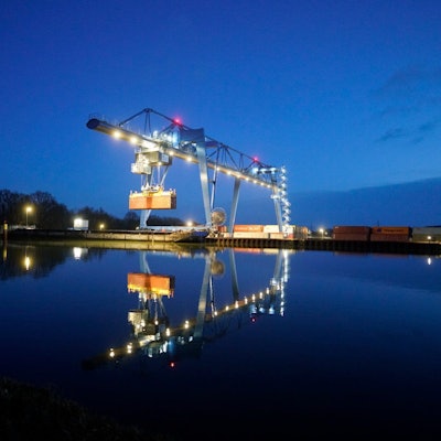 Im Wasser spiegelt sich ein beleuchteter Containerkran, der einen Container angehoben hat. (Archivbild)