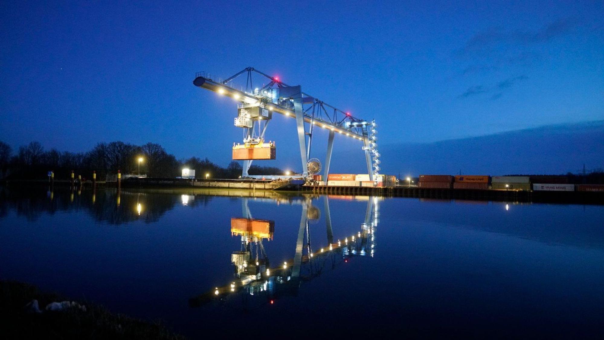 Im Wasser spiegelt sich ein beleuchteter Containerkran, der einen Container angehoben hat. (Archivbild)