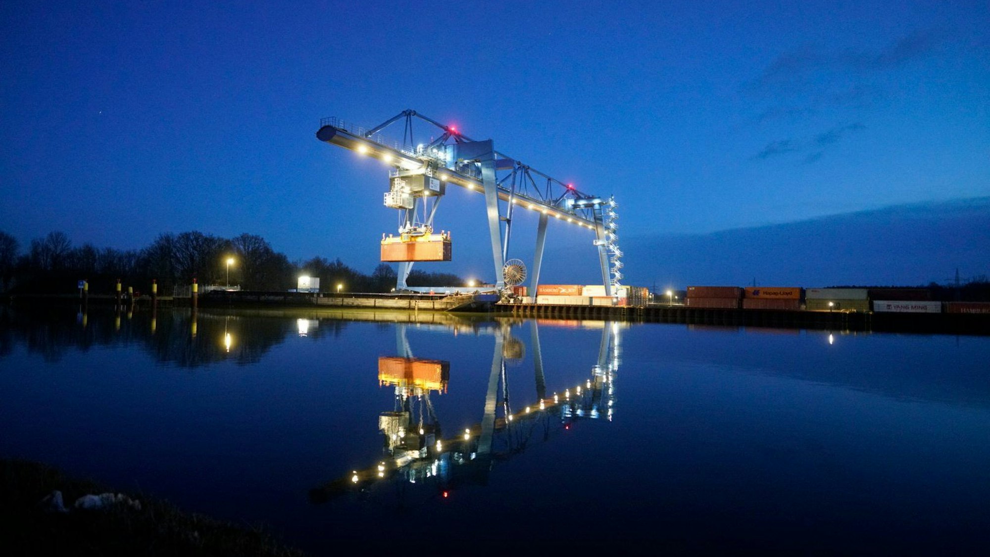 Im Wasser spiegelt sich ein beleuchteter Containerkran, der einen Container angehoben hat. (Archivbild)