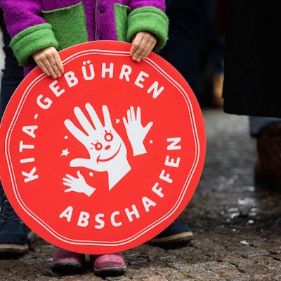 Ein Kind hält ein rotes, rundes Plakat mit der Aufschrift "Kita-Gebühren abschaffen" vor sich.