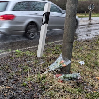 Am Straßenrand wurden an einem Baum Blumen und Kerzen abgelegt. Ein Auto fahrt vorbei.