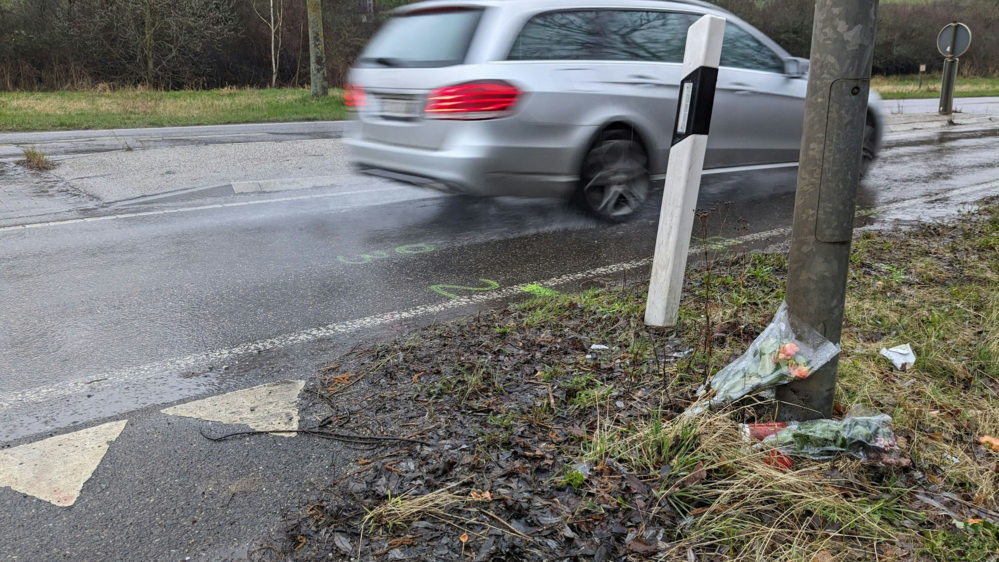 Am Straßenrand wurden an einem Baum Blumen und Kerzen abgelegt. Ein Auto fahrt vorbei.