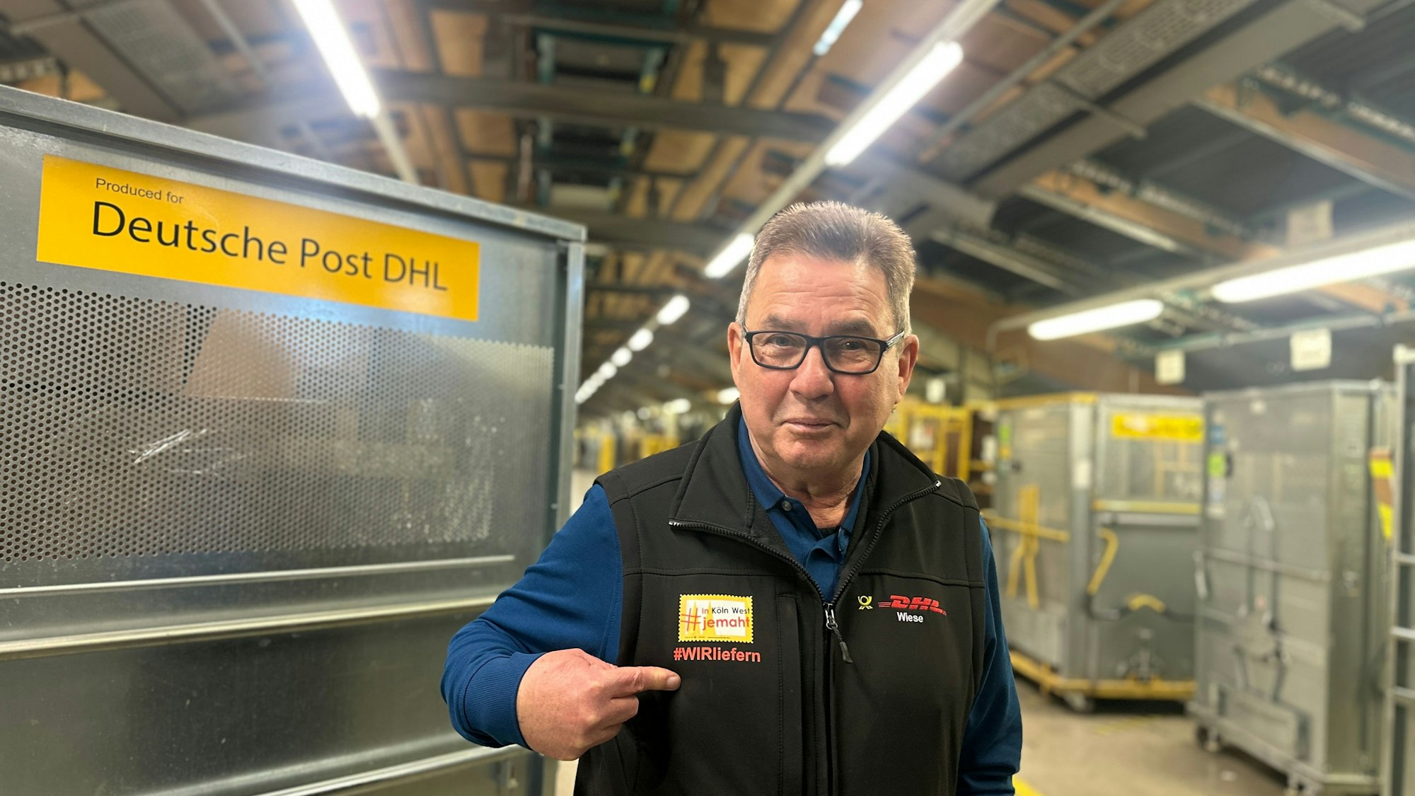 Ein älterer Mann mit Brille steht in einer Paketlagerhalle der Deutschen Post.