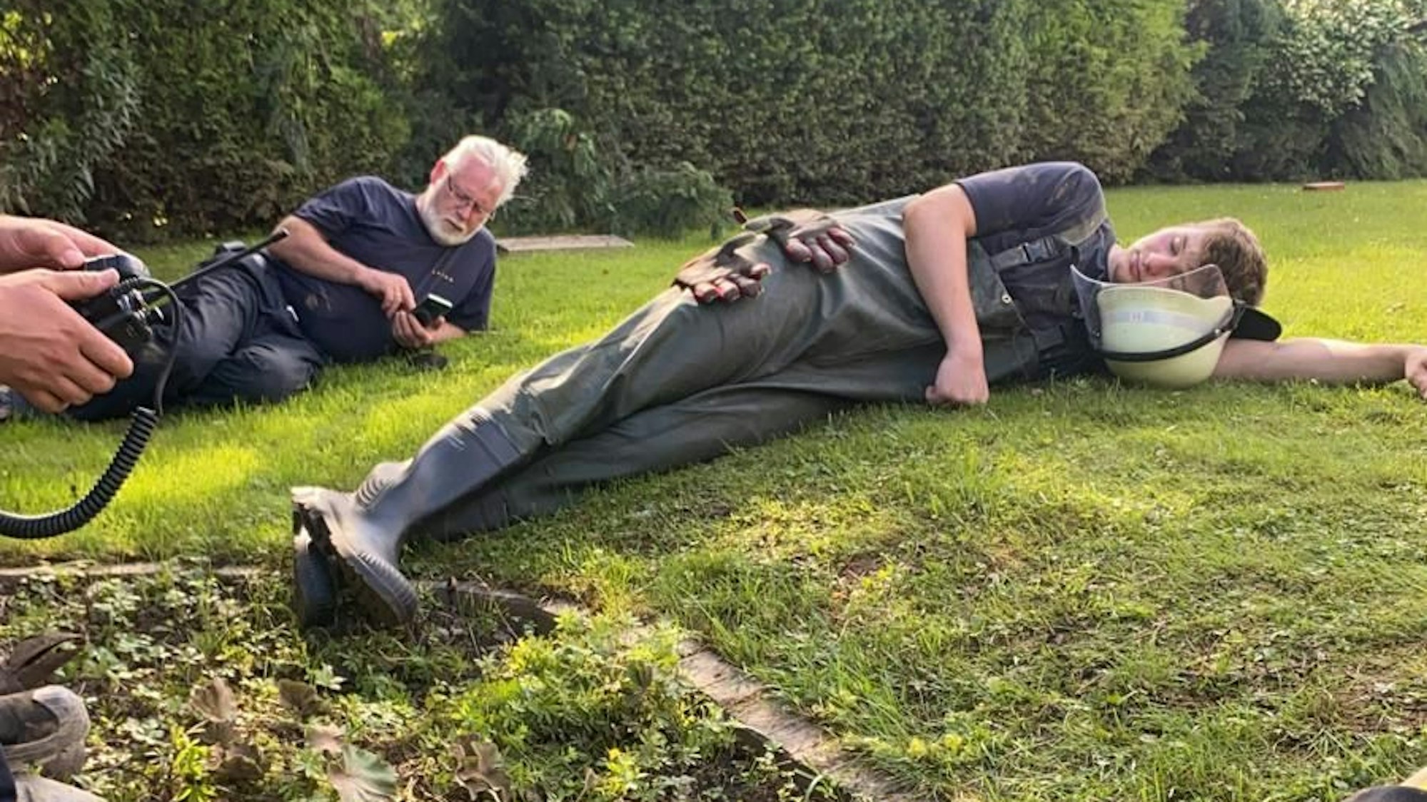 Das Bild zeigt eine junge Einsatzkraft, die auf der Wiese liegt und sich ausruht. Im Hintergrund schaut ein Feuerwehrmann in sein Handy.