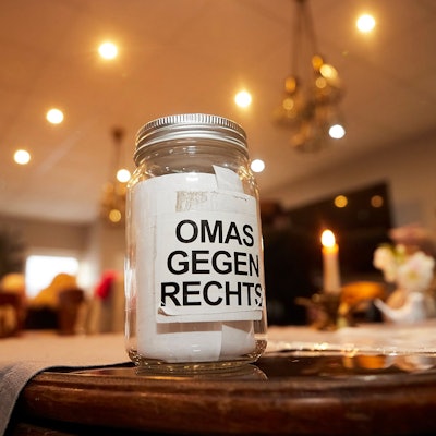 Ein Glas mit der Aufschrift Omas gegen Rechts steht auf einem Tisch. Im Hintergrund sind eine brennende Kerze und Menschen an weiteren Tischen zu erahnen.
