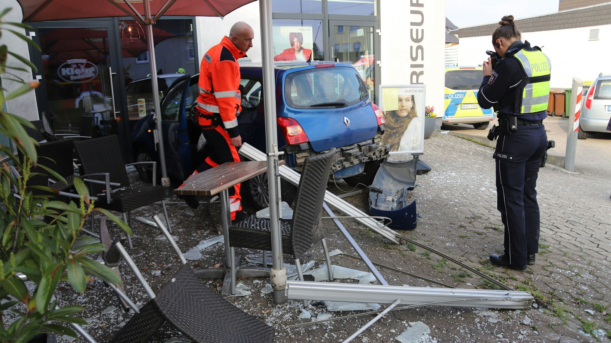 Ein Polizist fotografiert einen Kleinwagen, der in den Trümmern einer Außengastronomie steht, Glasscherben und zerstörte Möbel liegen auf dem Pflaster.