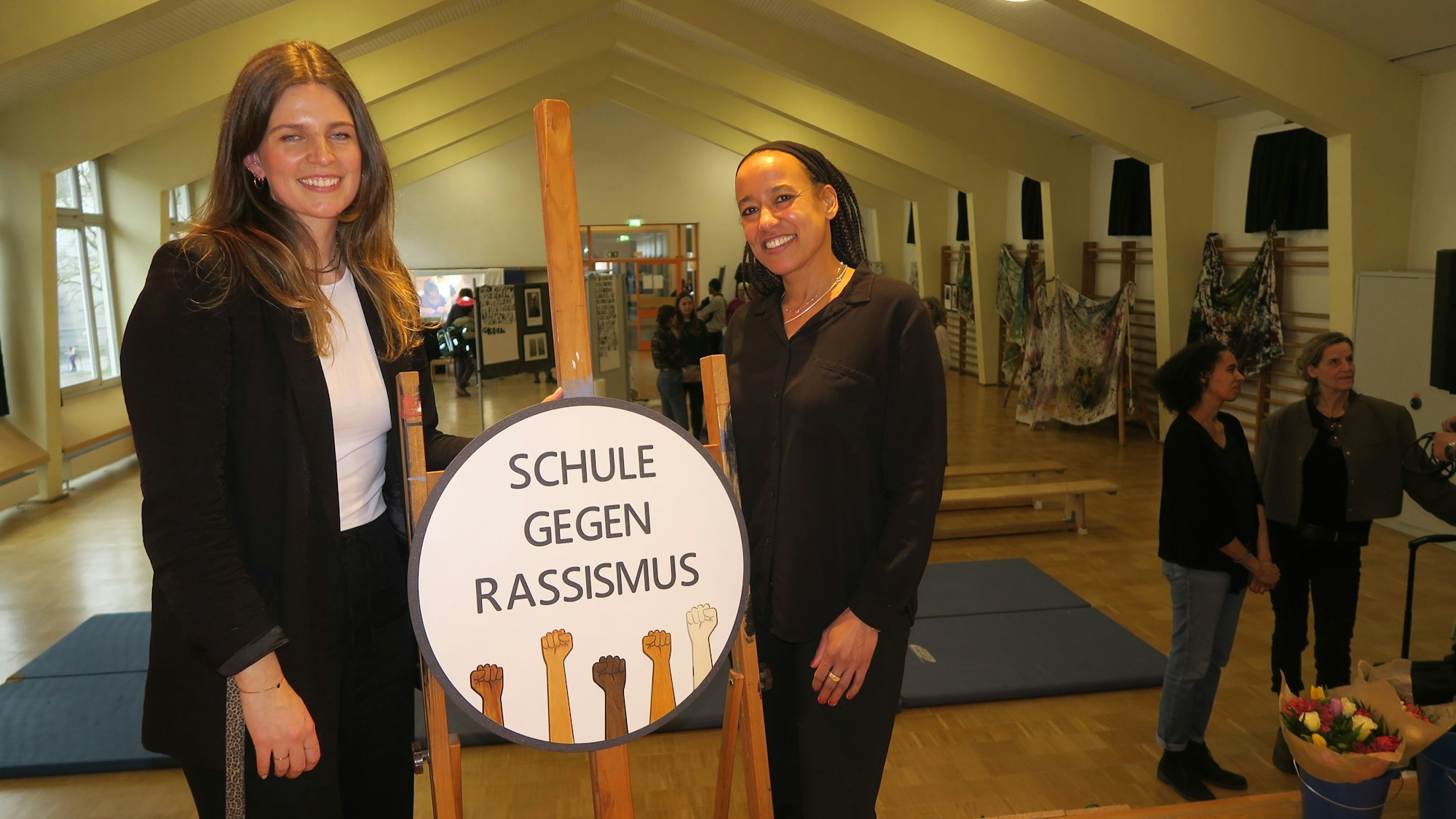In einer Turnhalle stehen zwei junge Frauen bei einem Aufsteller, auf dem Schule gegen Rassismus steht