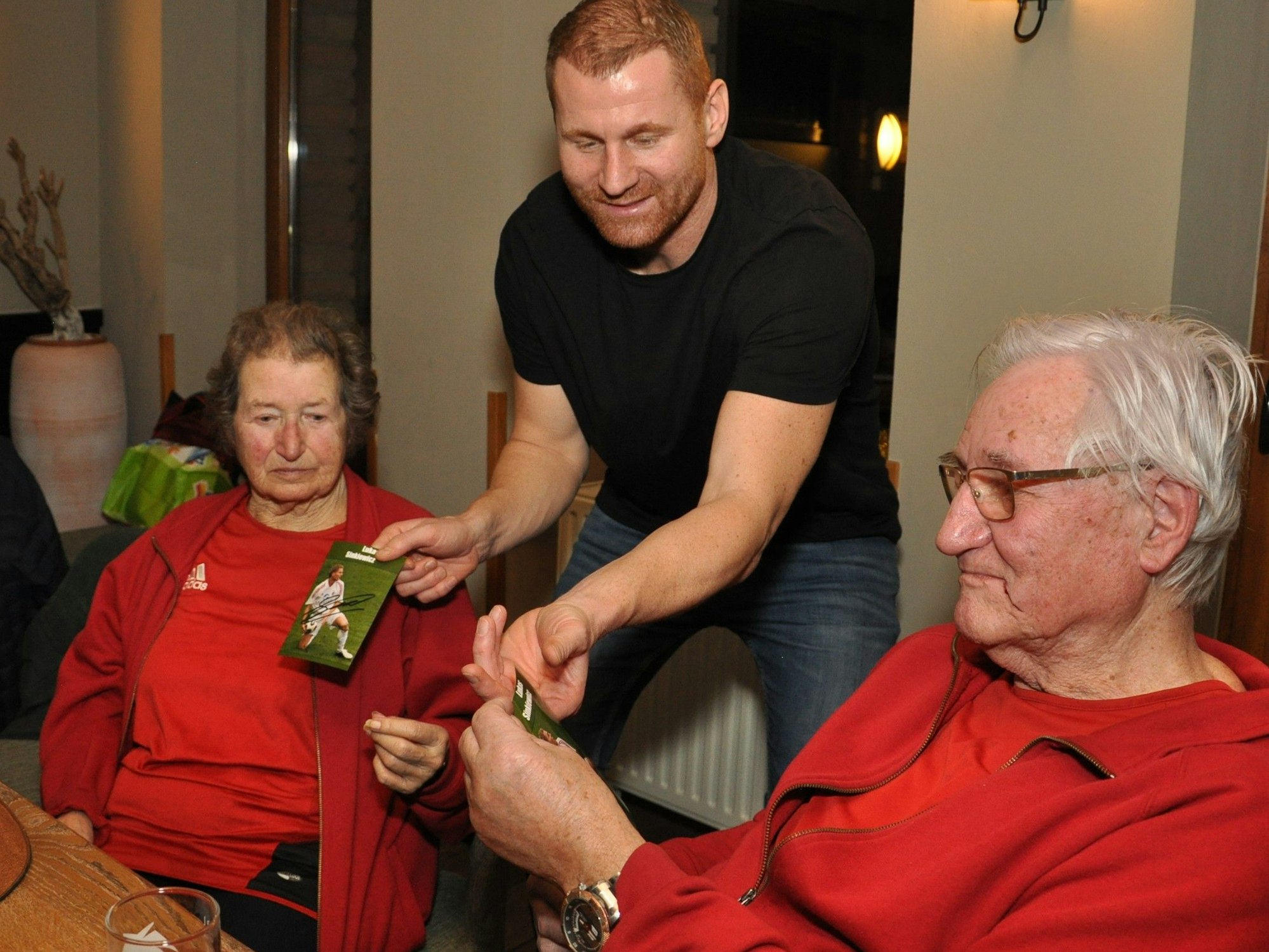 Ex-Nationalspieler Lukas Sinkiewicz steht in der Mitte des Bildes und überreicht einem am Tisch sitzenden Senioren-Ehepaar Autogrammkarten.