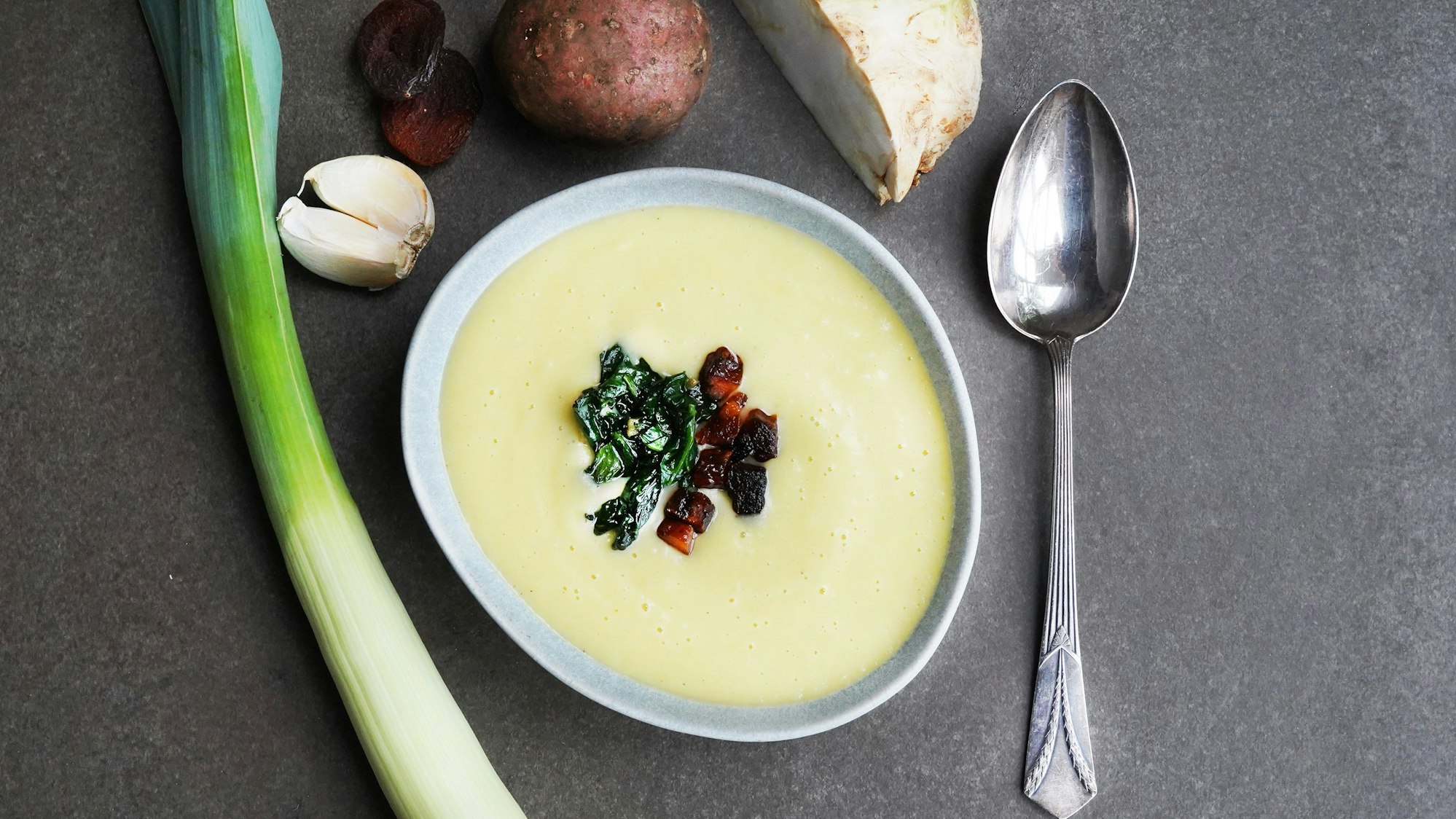 Die Suppe ist mit Spinat verziert in einem Suppenteller angerichtet. Daneben liegt ein Löffel und Gemüse.