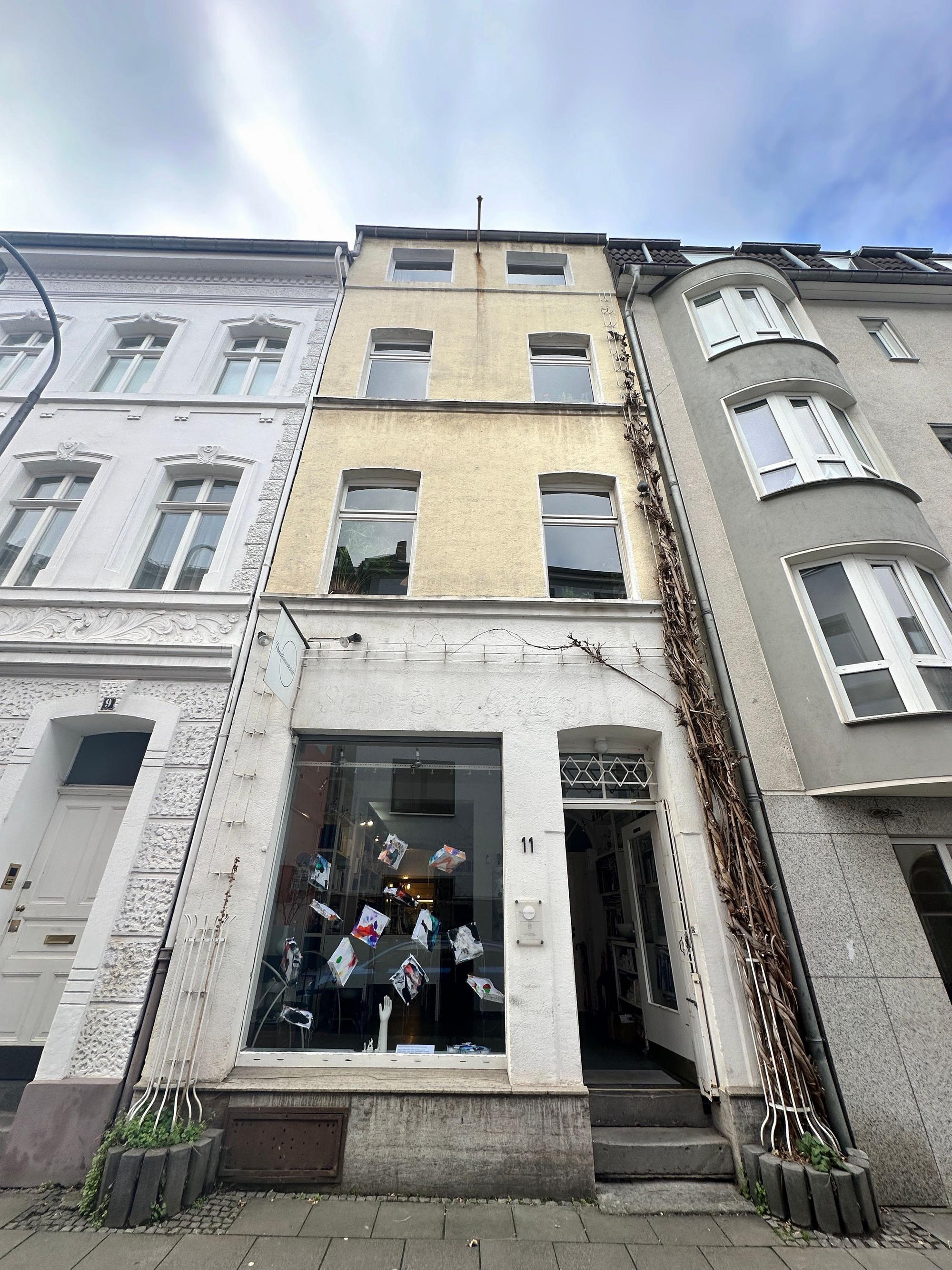 Blick auf ein schmales Haus mit zwei Fenstern pro Etage und einem Ladenlokal im Erdgeschoss