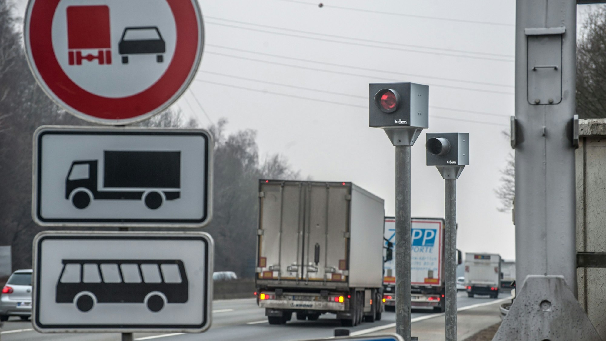 Ein Blitzer steht an einer Autobahn. An ihm fahren Lkws und Pkws vorbei. Vorne links steht ein Straßenschild.