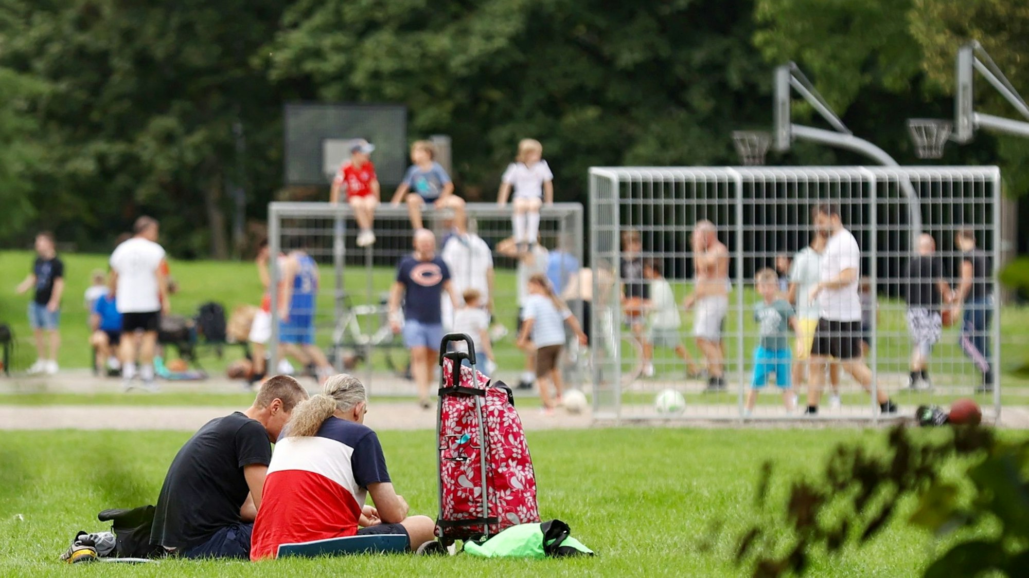 Viele Kinder und Jugendliche sitzen auf einer Wiese, im Hintergrund spielen einige Fußball auf einem Bolzplatz.