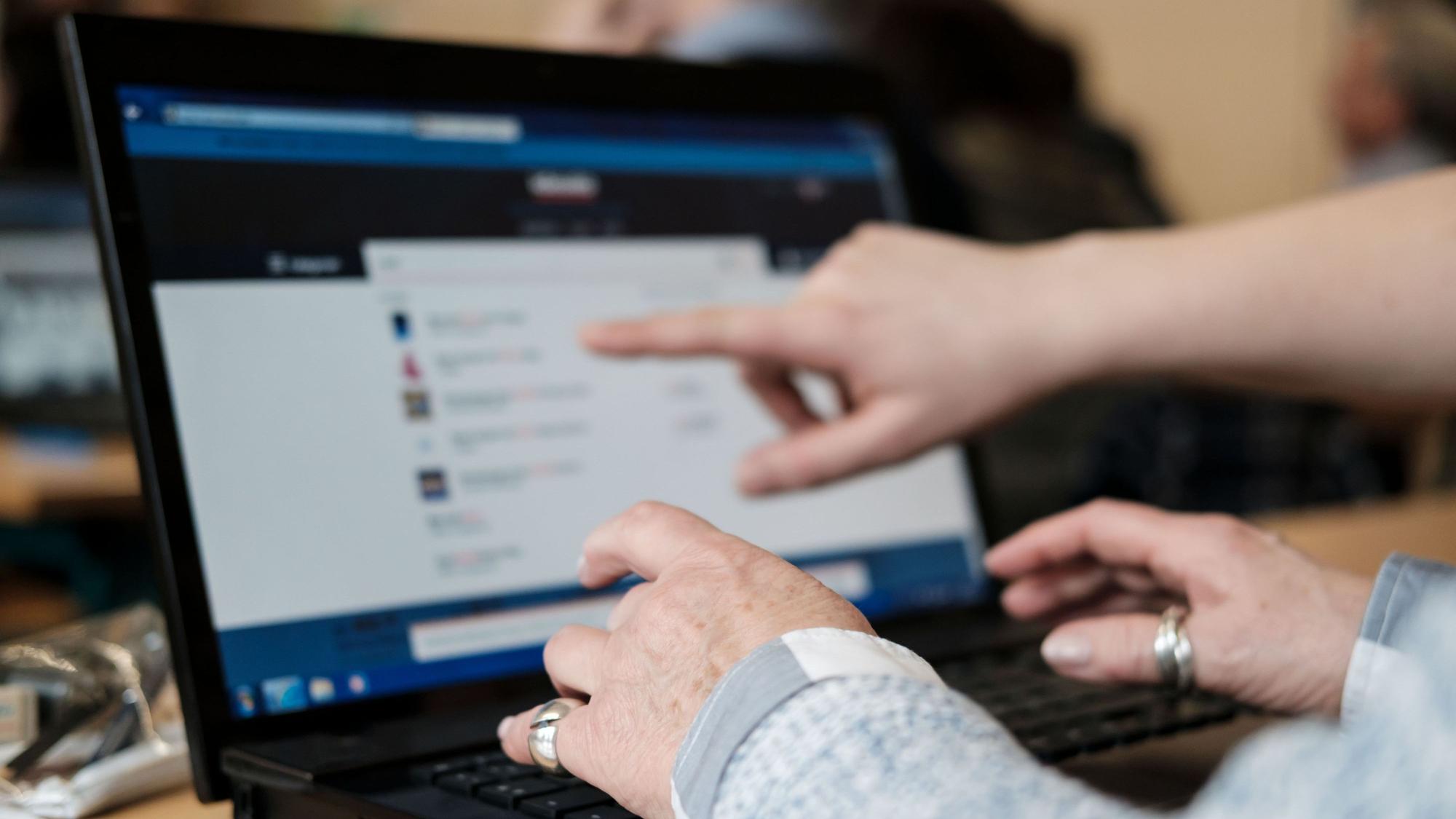 Eine Teilnehmerin des Computerkurses für Senioren übt zusammen mit einem Schüler an einem Laptop. Sie hält ihre Hände über die Tastatur, während ein Schüler mit einem Finger auf den Bildschirm zeigt.