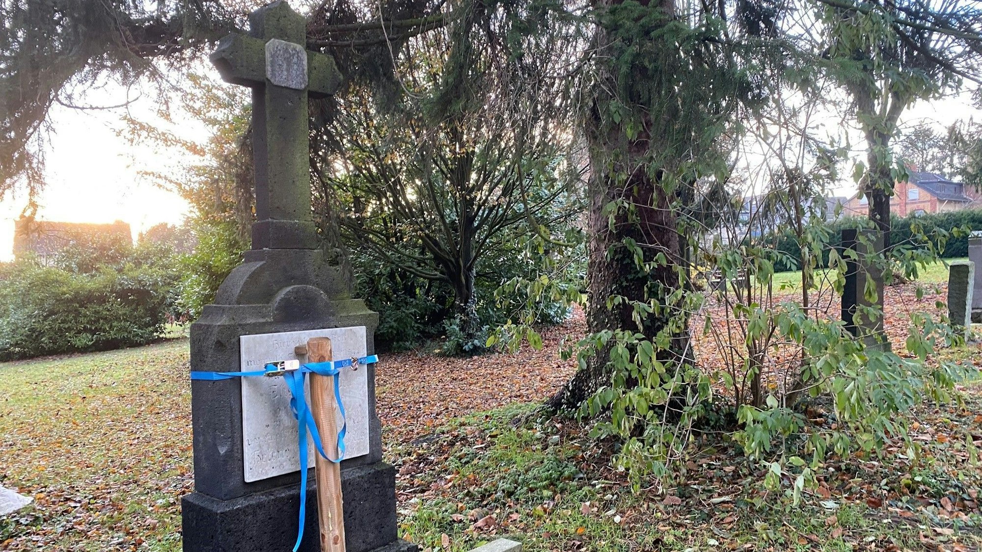 Zu sehen ist ein alter Grabstein, der mit einem Holzpflock und Riemen stabilisiert wurde.