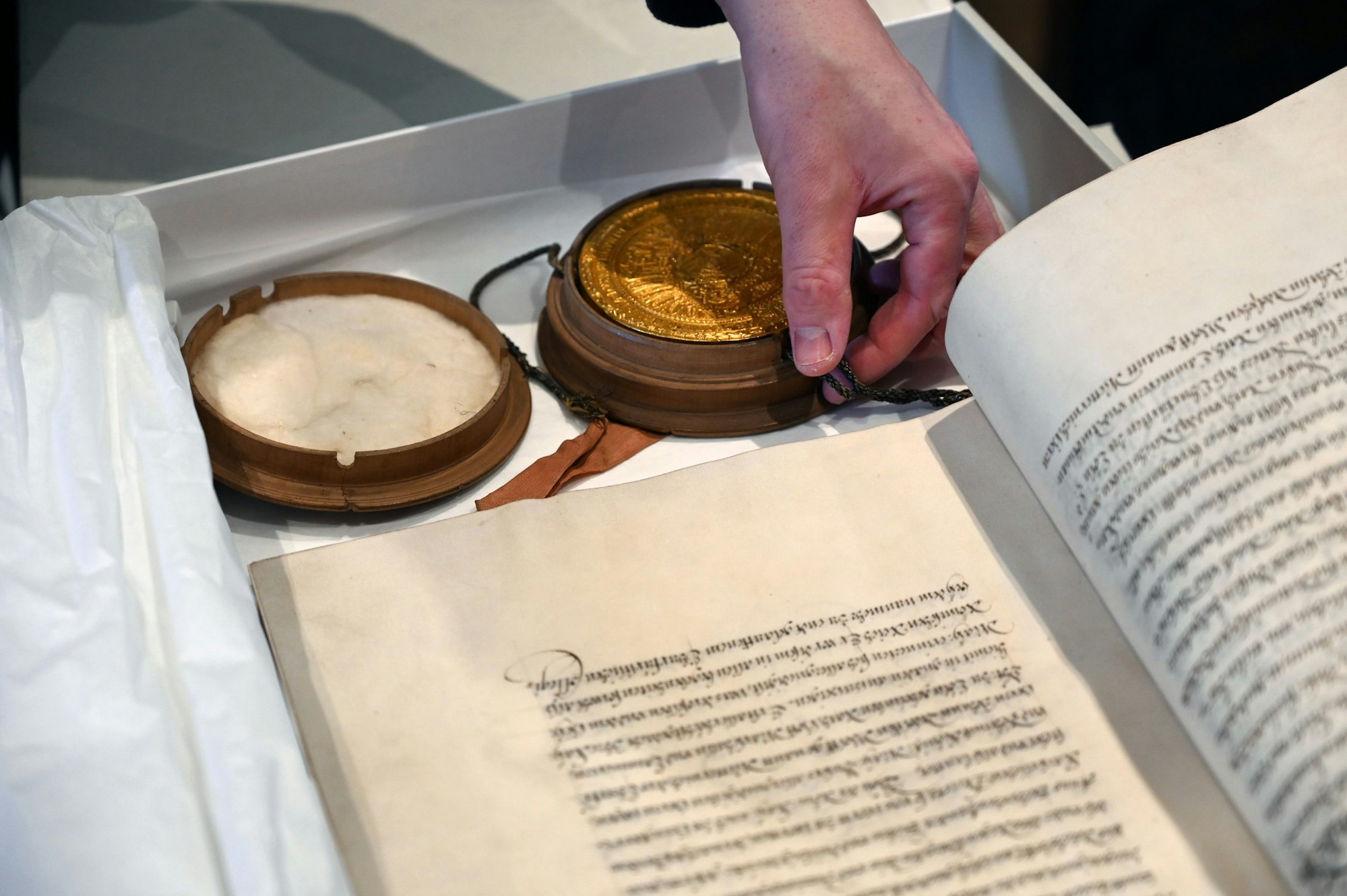 Eine Hand berührt ein Siegel in Gold unter einer mittelalterlichen Urkunde.