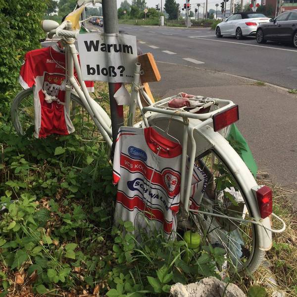Trikots hängen an einem weißen Fahrrad. Am Schildermast hängt ein Schild mit der Aufschrift „Warum bloß?“.