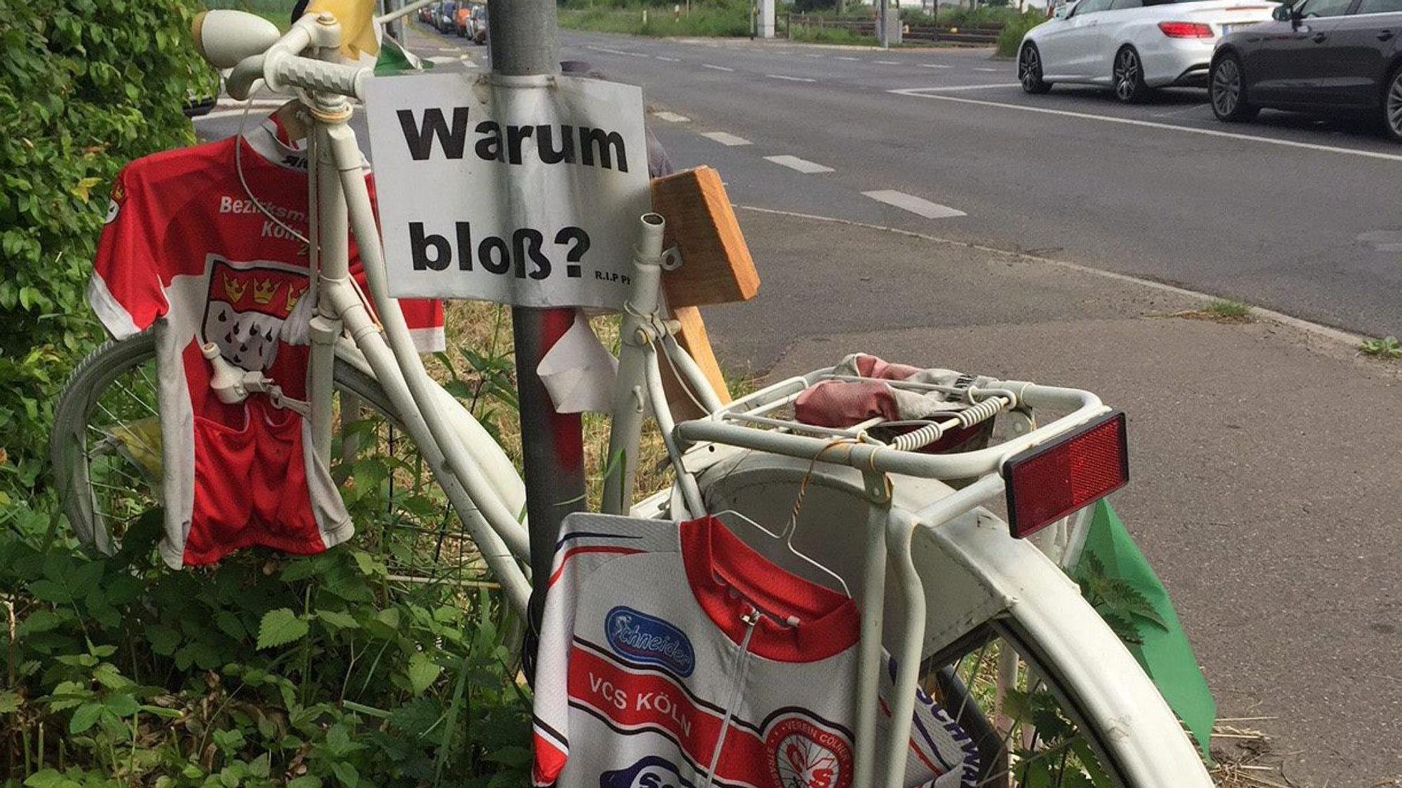 Trikots hängen an einem weißen Fahrrad. Am Schildermast hängt ein Schild mit der Aufschrift „Warum bloß?“.