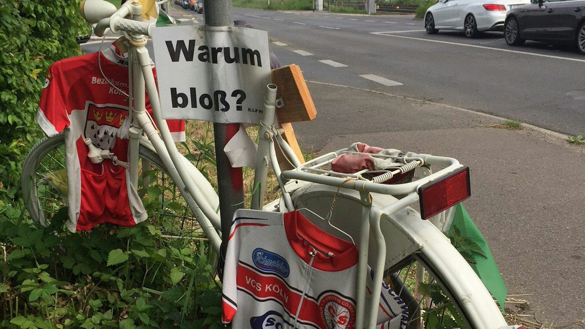 Ein weißes Fahrrad mit persönlichen Gegenständen wir Trikots steht in der Nähe der Unfallstelle am Straßenrand.