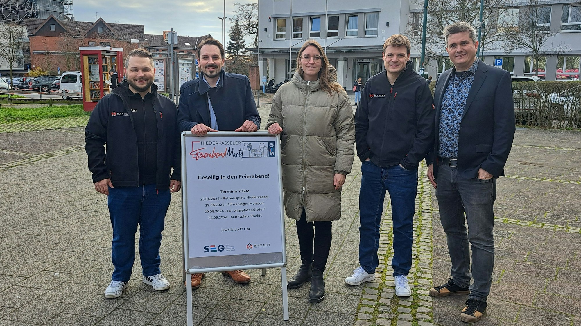 Die Organisatoren der geplanten Niederkasseler Feierabendmärkte stehen auf dem Rathausplatz und präsentieren das Plakat zur Veranstaltungsreihe.