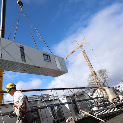 Ein rechteckiges Modul hängt an einem großen Baukran, im Vordergrund steht ein Bauarbeiter.