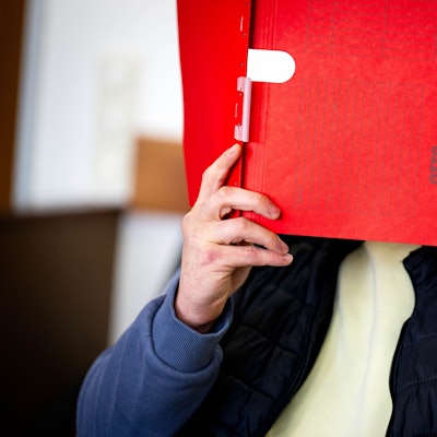 Ein Angeklagter hält sich im Gerichtssaal eine rote Mappe vor das Gesicht