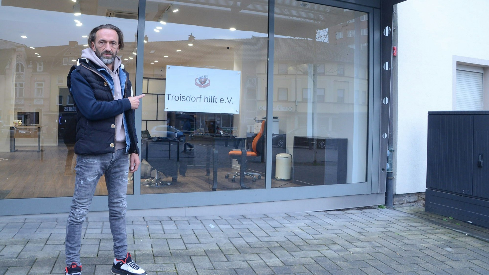 Ein Mann steht vor einem leeren Ladenlokal. Er zeigt auf ein Schild am Fenster mit der Aufschrift „Troisdorf hilft“.