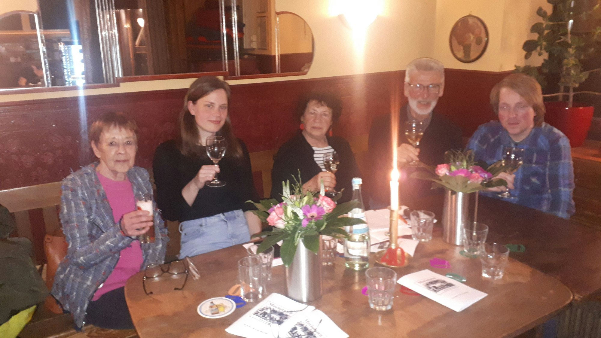 Fünf Personen sitzen mit erhobenen Gläsern an einem Tisch in einer Weinstube.