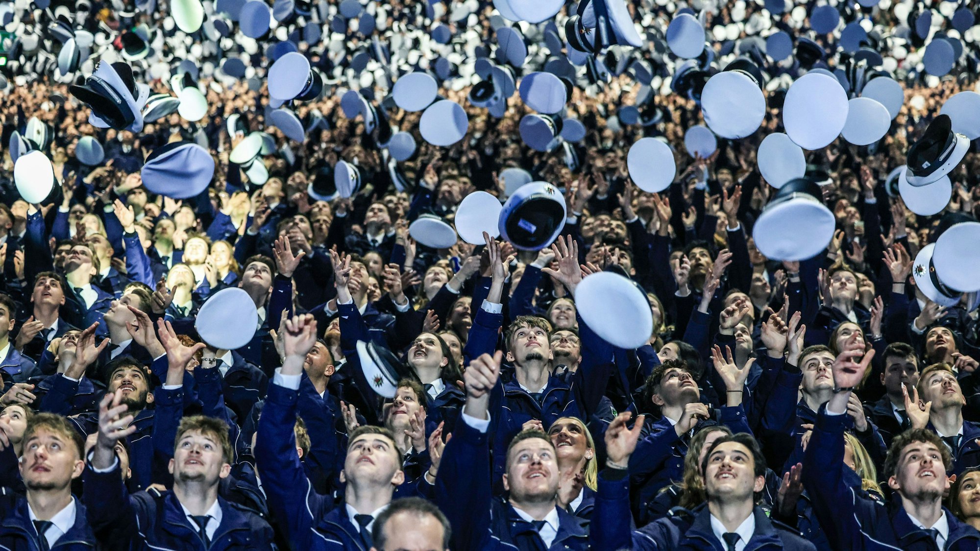 Köln: Polizei-Anwärter werfen am Ende der Vereidigungsfeier ihr Mützen hoch. In der Lanxess-Arena sind, teilweise von blauem Licht angestrahlt, mehr als 3000 angehende Polizistinnen und Polizisten vereidigt worden.