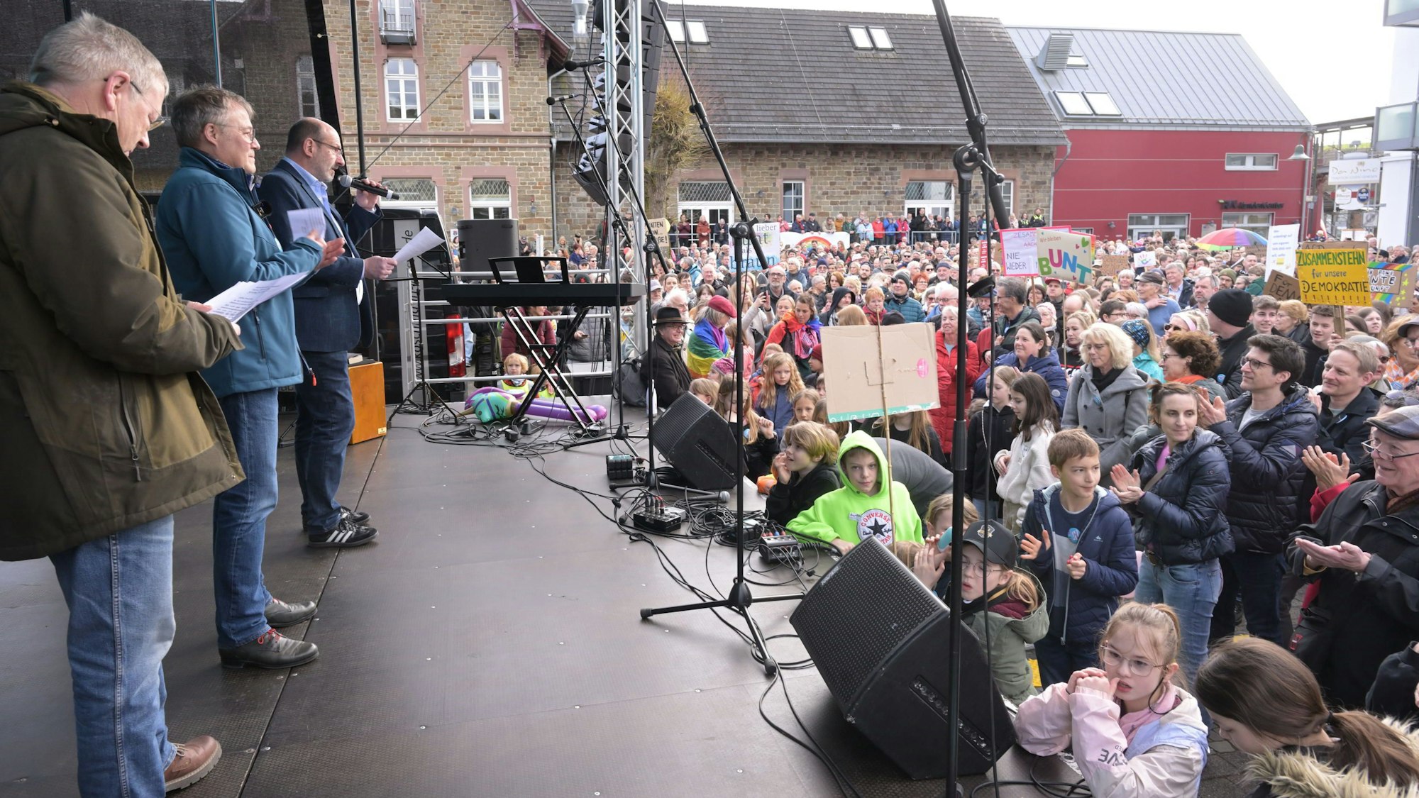 Drei Männer stehen auf einer Bühne vor Demonstrierenden auf dem Overather Bahnhofplatz.