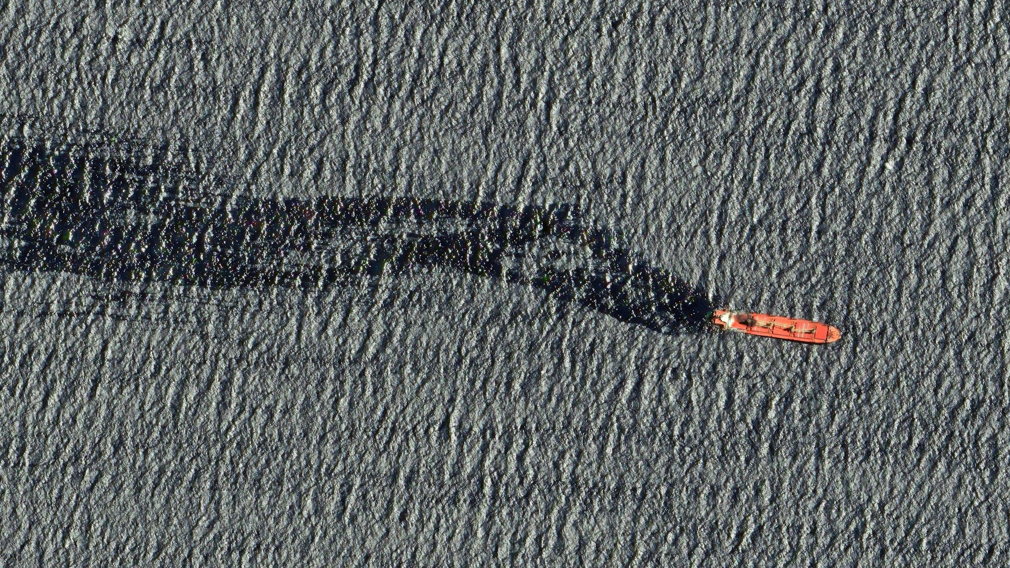 Bild von oben: Ein rotes Schiff fährt auf dem Meer. Der Frachter hinterlässt einen großen schwarzen Ölteppich hinter sich.