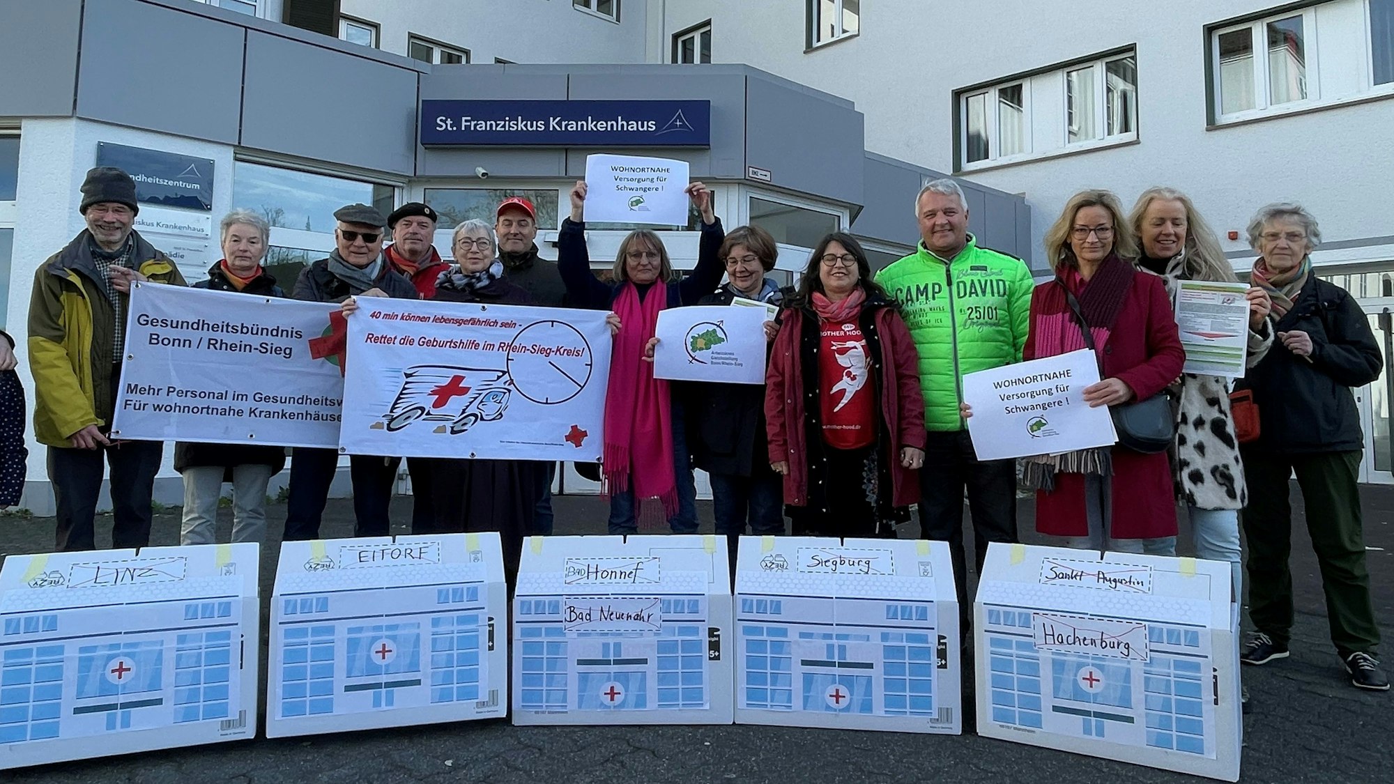 Menschen stehen vor dem Eingang des St. Franziskus-Krankenhaus Eitorf, vor ihnen Häuschen mit den Ortsnamen der bereits geschlossenen Geburtsstationen in der Region.