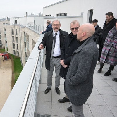 Auf der Dachterrasse stehen Bauherr, Investor und Bürgermeister und unterhalten sich. Zu sehen sind weitere im Viereck platzierte Gebäude. Im Hof ist bereits der Rasen verlegt.