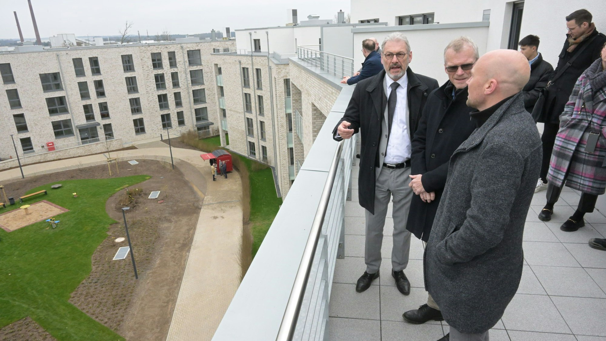 Auf der Dachterrasse stehen Bauherr, Investor und Bürgermeister und unterhalten sich. Zu sehen sind weitere im Viereck platzierte Gebäude. Im Hof ist bereits der Rasen verlegt.