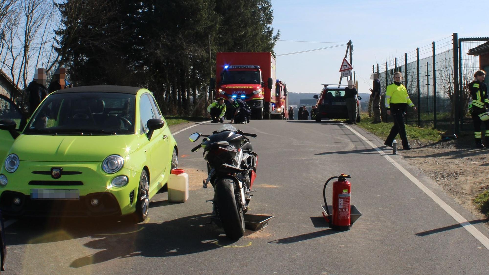 Beschädigte Zweiräder und ein Auto stehen auf einer Fahrbahn, auf dem Boden ist Ölbindemittel zu sehen. Im Hintergrund stehen Einsatzkräfte der Feuerwehr.