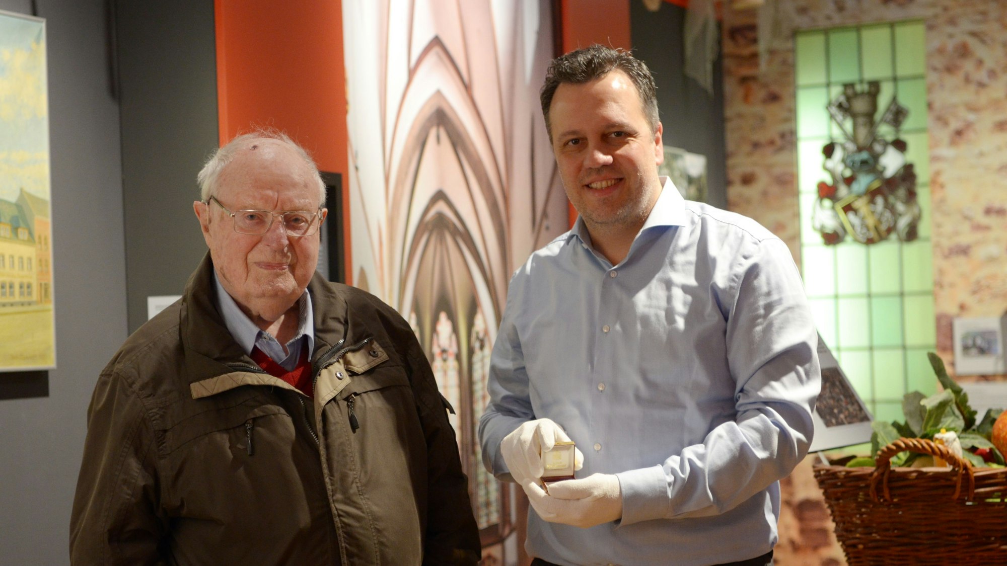 Rolf Müller-Blagovich und Sacha Reichelt stehen im Stadtmuseum Euskirchen. Reichelt zeigt eine der Münzen, die Müller der Stadt geschenkt hat.