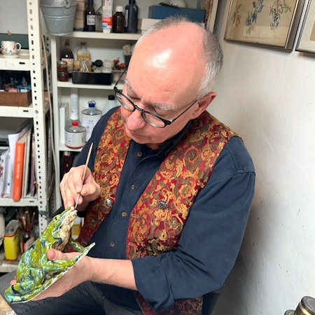 Ein Mann mit Glatze und Brille sitzt mit Schürze an einem Arbeitstisch und hantiert mit einer Porzellanfigur.