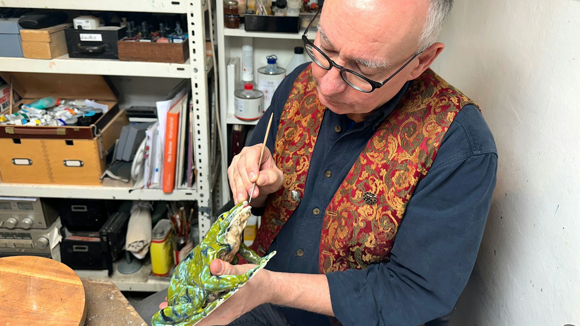 Ein Mann mit Glatze und Brille sitzt mit Schürze an einem Arbeitstisch und hantiert mit einer Porzellanfigur.