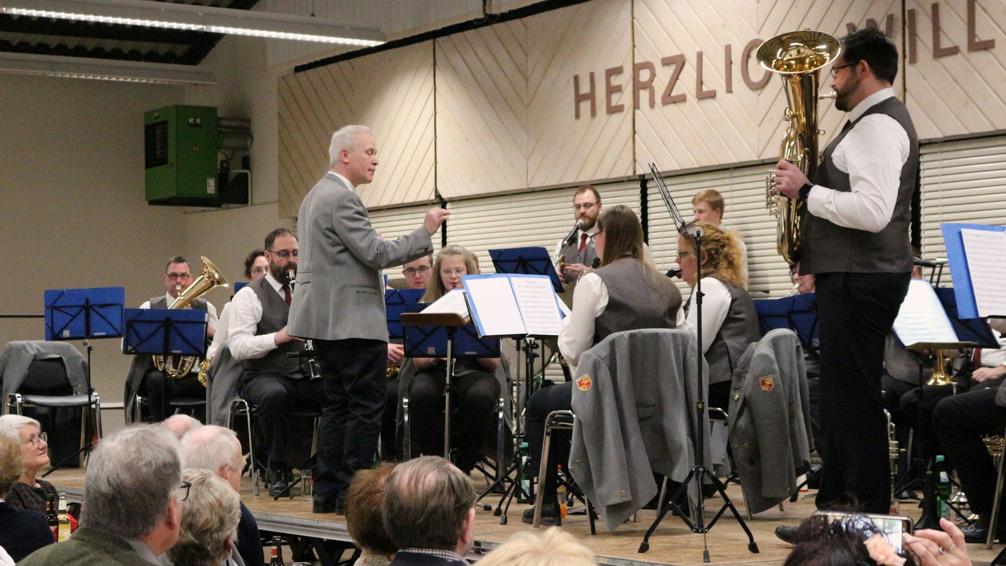 Das Orchester spielt auf der Bühne der Schwerfener Schützenhalle, dirigiert von Rudi Wedel. Im Vordergrund sieht man Teile des Publikums.