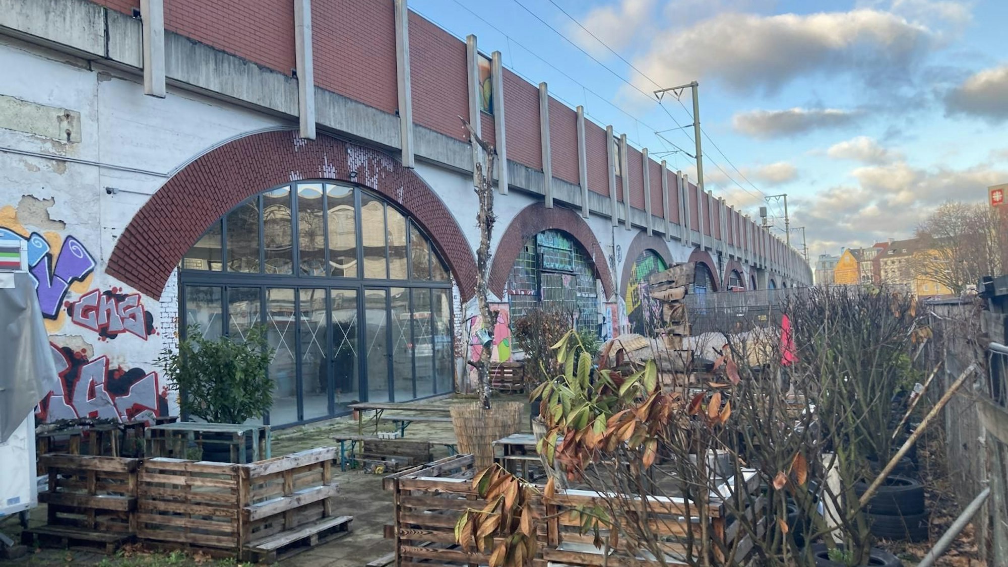 Vor dem Bahndamm mit verglasten Bahnbögen blickt man auf winterlich-verwelkte Pflanzen, die eine Biergarten-Fläche umranden.