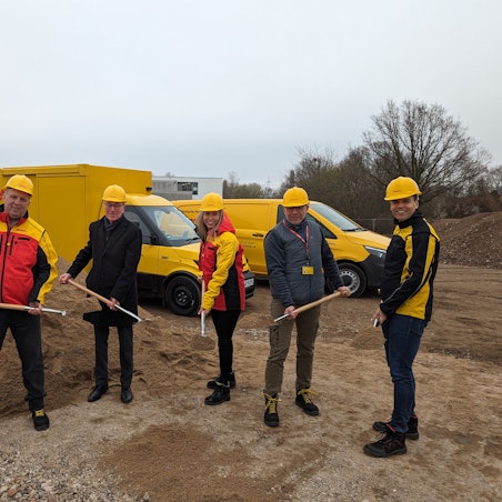 Vier Männer und eine Frau mit gelben Helmen und Spaten auf der Baustelle. Im Hintergrund gelbe Zustellfahrzeuge der Post.
