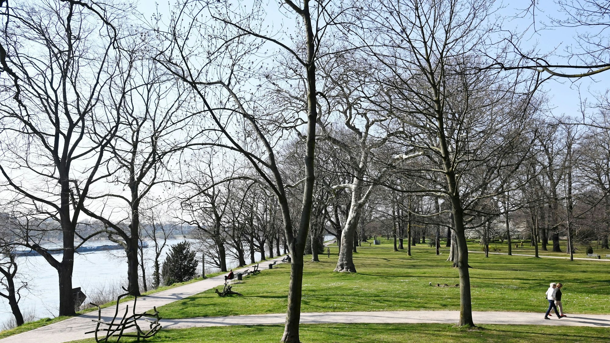 Auf dem Bild ist der Park zu sehen, auf den Wiesen einige Bäume und Skulpturen. Rechts gehen zwei Menschen einen Weg entlang, links im Bild sieht man das Rheinufer.