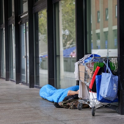 Das Bild zeigt einen Obdachlosen in einem Schlafsack, der neben sich seine Habseligkeiten in einem Einkaufswagen aufbewahrt.
