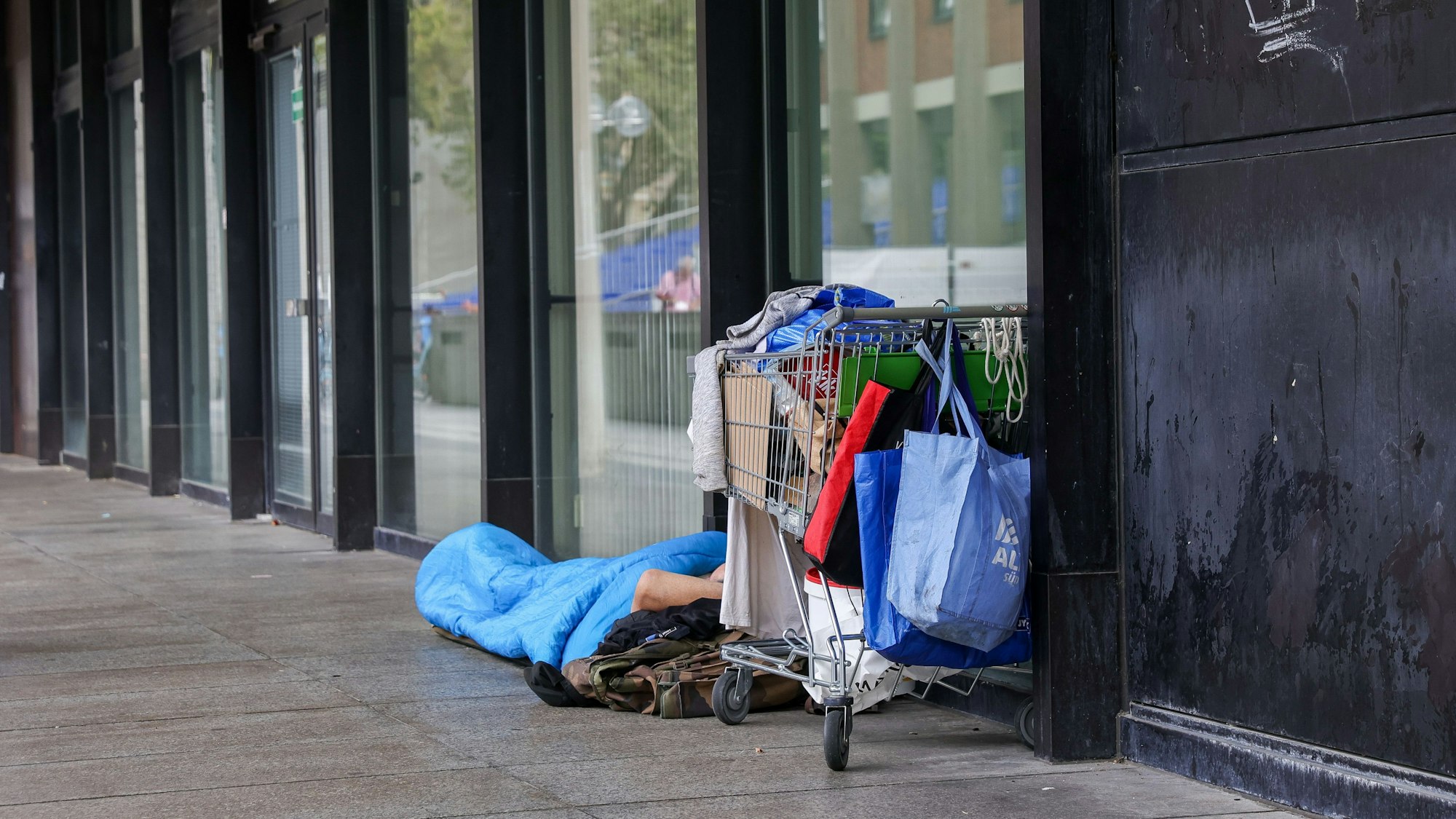 Das Bild zeigt einen Obdachlosen in einem Schlafsack, der neben sich seine Habseligkeiten in einem Einkaufswagen aufbewahrt.